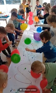 Dzieci stoją dookoła długiego arkusza białego papieru. W rączkach trzymają balony na sznurkach , którymi stemplują kolorową farbkę, rozmieszczoną na arkuszu.