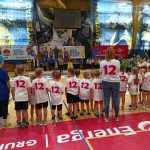 Grupa dzieci wraz z panią stoją tyłem do zdjęcia, mają na sobie białe koszulki z krótkim rękawem i krótkie czarne spodenki. Na plecach widnieje liczba 12 w kolorze czerwonym. W tle widać duże okna i reklamy, które znajdują się w hali sportowej. Na pierwszym planie duży napis ENERGA na czerwonym tle.