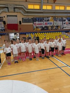 Grupa dzieci pozuje do zdjęcia, tym razem stoją przodem. Uśmiechają się. Mają na sobie strój sportowy - biała koszulka z krótkim rękawkiem i logo przedszkola Tęcza oraz czarne, krótkie spodenki. W tle widać trybuny hali sportowej. tzn żółte krzesełka.