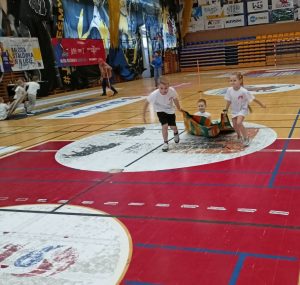 Dwoje dzieci (chłopiec i dziewczynka) ciągną chłopca siedzącego na kocu po podłodze hali sportowej szkoły podstawowej. Dzieci mają na sobie białe koszulki sportowe z krótkim rękawem oraz czarne, krótkie spodenki.