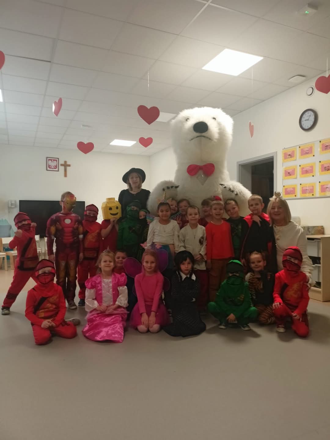 Grupa dzieci w wieku przedszkolnym pozuje do zdjęcia w przebraniach z okazji karnawału. Każde dziecko ma na sobie inne przebranie. Na środku stoi wielki, biały niedźwiedź - osoba przebrana za misia, która była animatorem i zabawiała dzieci.