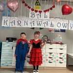 Dwoje dzieci - chłopiec i dziewczynka pozują do zdjęcia na tle napisu BAL KARNAWAŁOWY. Dziewczynka jest przebrana za Hiszpankę/ tancerkę a chłopiec za Supermana. Dzieci są wesołe i radosne. Z sufitu zwisają balony i serduszka.