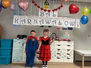 Dwoje dzieci - chłopiec i dziewczynka pozują do zdjęcia na tle napisu BAL KARNAWAŁOWY. Dziewczynka jest przebrana za Hiszpankę/ tancerkę a chłopiec za Supermana. Dzieci są wesołe i radosne. Z sufitu zwisają balony i serduszka.