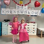Dwie dziewczynki w różowych strojach księżniczek pozują na tle z napisem BAL KARNAWAŁOWY. Z sufitu zwisają balony i serduszka.