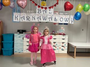 Dwie dziewczynki w różowych strojach księżniczek pozują na tle z napisem BAL KARNAWAŁOWY. Z sufitu zwisają balony i serduszka.
