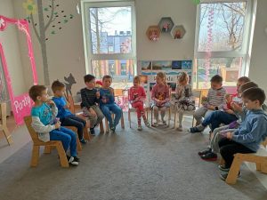 Grupa dzieci w wieku przedszkolnym siedzi w sali na krzesełkach. Na pierwszym planie widać duży, szary dywan. Trzy dziewczynki siedzą na środku, ponieważ dziś jest ich święto - dzień kobiet. Chłopcy wręczyli im prezenty - torebki i żelki. W tle widać duże okna oraz plac zabaw.