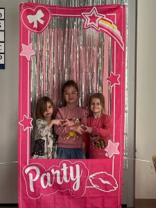 Trzy wesołe dziewczynki ubrane w sukienkę stoi w foto budce z okazji Dnia Kobiet. Jest to różowa ramka imitująca ramkę do zdjęcia, w kolorze różowym, z gwiazdkami. W tle jest srebrna serpentyna.