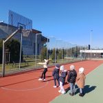 Pięć dziewczynek stoi na boisku, jedna rzuca piłką do kosza.