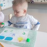 Dziecko siedzi przy białym stoliku, trzyma w ręce kolorowy pisak, którym rysuje, kółka, kreski w konturach garnka.
