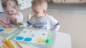 Dziecko siedzi przy białym stoliku, trzyma w ręce kolorowy pisak, którym rysuje, kółka, kreski w konturach garnka.
