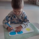Dziecko siedzi przy białym stoliku, trzyma w ręce kolorowy pisak, którym rysuje, kółka, kreski w konturach garnka.