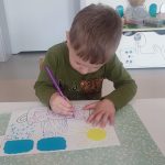 Dziecko siedzi przy białym stoliku, trzyma w ręce kolorowy pisak, którym rysuje, kółka, kreski w konturach garnka.