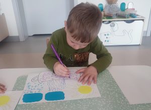 Dziecko siedzi przy białym stoliku, trzyma w ręce kolorowy pisak, którym rysuje, kółka, kreski w konturach garnka.