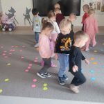 Duża grupa dzieci chodzi po szarym dywanie, na którym rozłożone są kwiatki kolorowe.