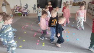 Duża grupa dzieci chodzi po szarym dywanie, na którym rozłożone są kwiatki kolorowe.