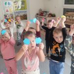 Dzieci trzymają do góry ręce, w których trzymają dwa kolorowe kwiatki.