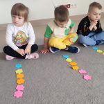 Troje dzieci siedzi na szarym dywanie. Przed nimi leżą ułożone w kolumnie kolorowe kwiatki. Jest ich sześć.