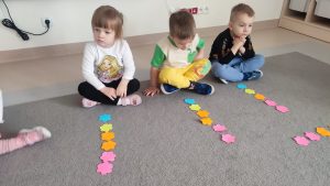 Troje dzieci siedzi na szarym dywanie. Przed nimi leżą ułożone w kolumnie kolorowe kwiatki. Jest ich sześć.