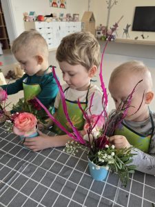 Trzech chłopców siedzi przy szarym stole i tworzy bukiety z kolorowych ciętych kwiatów.