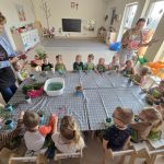 Duża grupa dzieci siedzi na około kwadratowego dużego stołu przykrytego szarą ceratą. Na stole leżą kwiaty miska z wodą. Przy dzieciach stoją dwie kobiety.