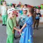Dziewczynka w niebieskiej sukience w kwiaty, tańczy razem z trzema chłopcami z czapkami bocianów na głowach.