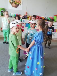 Dziewczynka w niebieskiej sukience w kwiaty, tańczy razem z trzema chłopcami z czapkami bocianów na głowach.