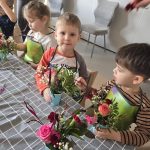 Dzieci ubrane w zielony fartuszek siedzą przy szarym stoliku. Przed nimi bukiety z żywych kwiatów.