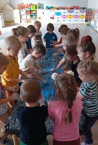 Grupa dzieci dookoła stolika , na którym jest położona czarna folia, a na niej niebieska farba, którą dzieci rozprowadzają rączkami