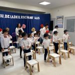 Dzieci stoją na sali za małymi krzesełkami pokazując układ taneczny.