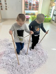 Dwóch chłopców ma założone na nogach "narty" i wjeżdżają w górkę śniegu.