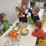 Dzieci z pomocą nauczyciela wkładają do sokowirówki owoce.