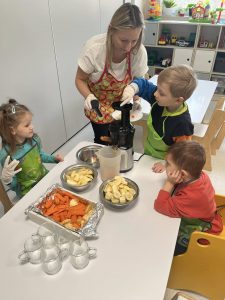 Dzieci z pomocą nauczyciela wkładają do sokowirówki owoce.
