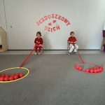 Dwoje dzieci siedzą na krzesełkach i przyciągają do siebie hula hop wypełnione piłeczkami w kolorze czerwonym.