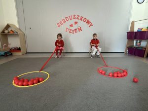 Dwoje dzieci siedzą na krzesełkach i przyciągają do siebie hula hop wypełnione piłeczkami w kolorze czerwonym.