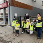 Dzieci z nauczycielami i opiekunami, ubrane w kamizelki stoją przed cukiernią.