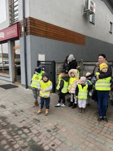 Dzieci z nauczycielami i opiekunami, ubrane w kamizelki stoją przed cukiernią.