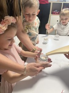 Dzieci z pomocą nauczyciela i opiekuna wlewają wodę do słoiczka z gazą.