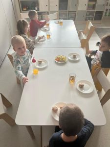 Dzieci siedzą przy stoliczkach i jedzą pączki.
