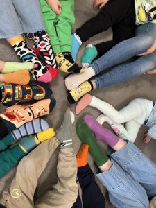 Dzieci siedzą w kole i pokazują eksponują swoje kolorowe skarpetki, które są symbolem solidarności z osobami z Zespołem Downa.
