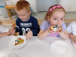 Dzieci siedzą przy stoliczku i degustują samodzielnie udekorowane gofry.