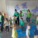 Dzieci tańczą w kole. Na dywanie jest rozłożona folia imitująca wodę, do której żabki będą wskakiwać.