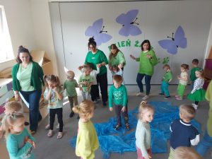 Dzieci tańczą w kole. Na dywanie jest rozłożona folia imitująca wodę, do której żabki będą wskakiwać.
