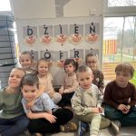 Grupka dzieci siedząc pozuje do zdjęcia na tle tablicy z napisem dzień gorra.