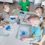 Dzieci siedzą przy białych stolikach i malują serduszko niebieską farbą.