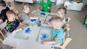 Dzieci siedzą przy białych stolikach i malują serduszko niebieską farbą.