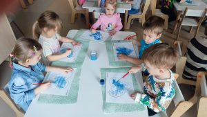Dzieci siedzą przy białych stolikach i malują serduszko niebieską farbą.