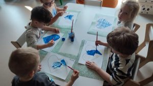 Dzieci siedzą przy białych stolikach i malują serduszko niebieską farbą.
