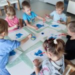 Dzieci siedzą przy białych stolikach i malują serduszko niebieską farbą.