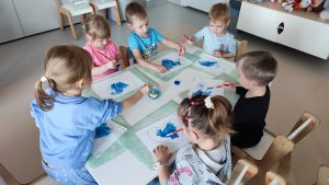 Dzieci siedzą przy białych stolikach i malują serduszko niebieską farbą.
