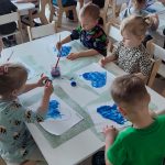 Dzieci siedzą przy białych stolikach i malują serduszko niebieską farbą.